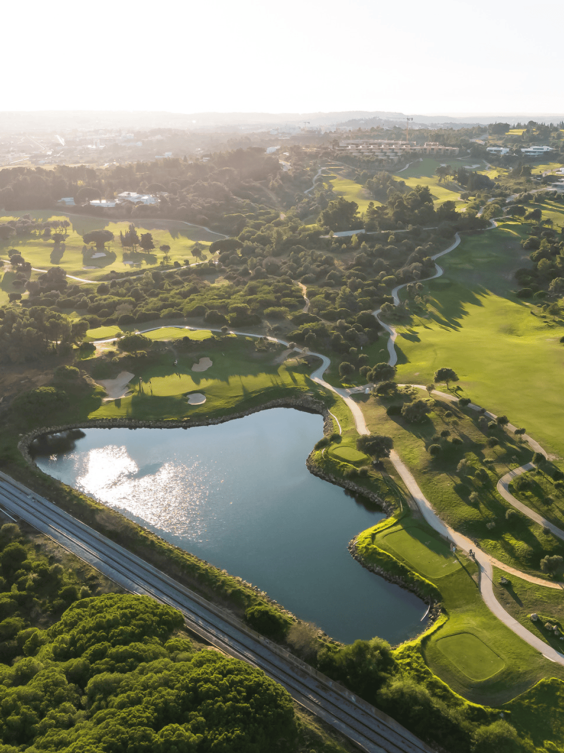 Vista aérea do campo de golfe de Palmares com lago em forma de coração, linha ferroviária e colinas verdes, captando a diversidade da paisagem envolvente.